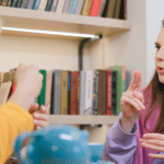 Two female students having an animated discussion in a library setting, surrounded by books.