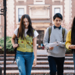 Three students walking and discussing with notebooks and books in hand in a university campus setting.